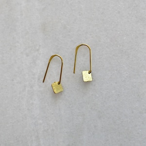 Op de afbeelding: Een paar goudkleurige oorbellen. Elke oorbel heeft een gebogen, langwerpige haak met een kleine, vierkante hanger. De oorbellen hebben een minimalistisch ontwerp en een getextureerd oppervlak.