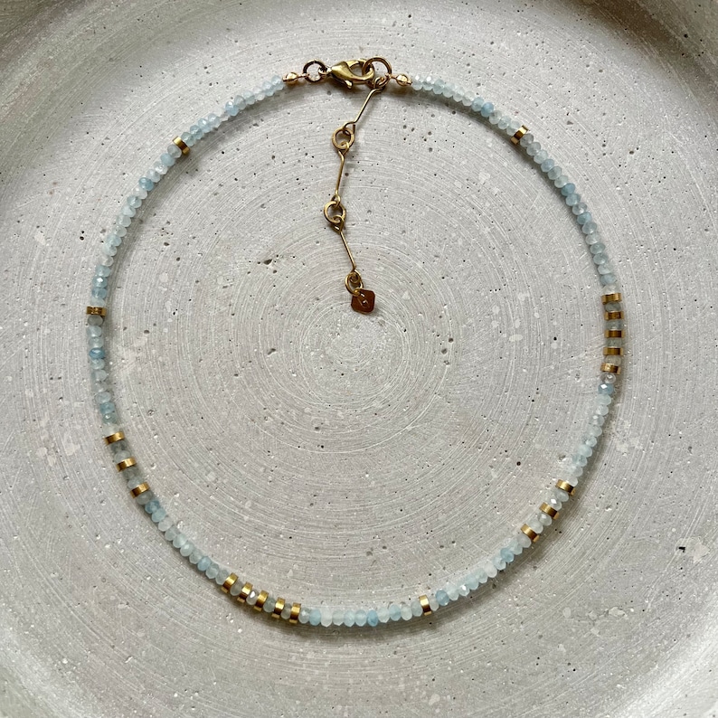 May include: A beaded necklace featuring small, faceted light blue stones interspersed with cylindrical gold-toned metal spacers. The necklace includes a gold-toned lobster clasp, an adjustable chain extension, and a small flat charm at the end.