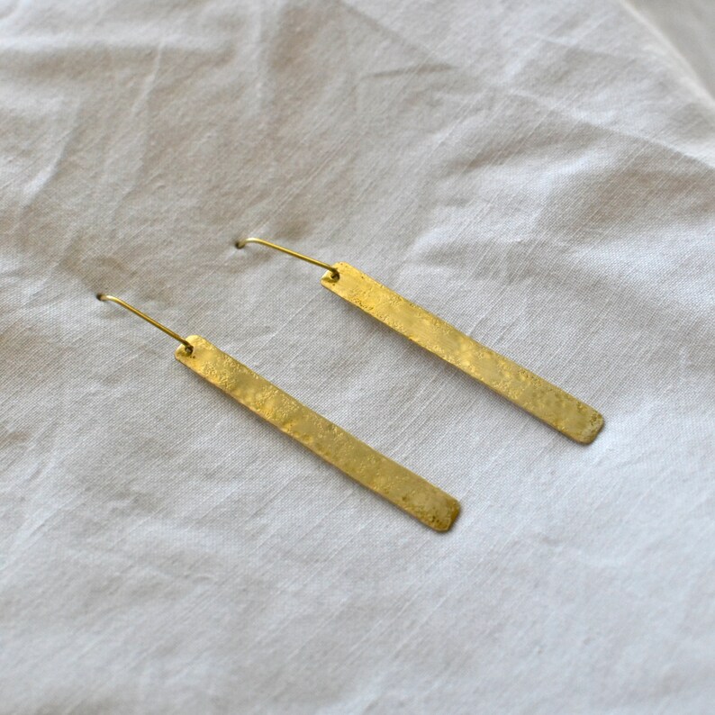 May include: A pair of gold-toned earrings featuring long, slender rectangular bars with a textured, hammered finish. Each earring is attached to a thin, curved wire hook designed for insertion through the earlobe, showcasing a metallic luster.