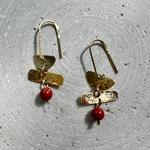 Könnte beinhalten: Ein Paar goldfarbene Ohrringe mit gehämmerten Strukturen. Jeder Ohrring hat eine halbmondförmige Form, einen rechteckigen Balken und eine kleine, runde, rote Perle. Die Ohrringe hängen an einem gebogenen Haken.
