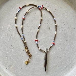 May include: A beaded necklace with a dark brown cord, featuring white, blue, and red beads. The necklace has a gold-colored clasp and a dark, elongated pendant. The beads are small and evenly spaced.