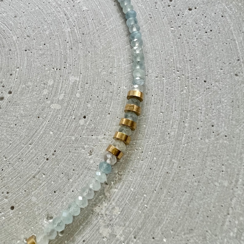 May include: A necklace strand featuring small, faceted, pale blue beads arranged in a curved line. Interspersed among the blue beads are several cylindrical, metallic gold-toned spacers that create a distinct contrast in texture and color.