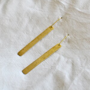 May include: A pair of long, thin, rectangular brass earrings with a textured, matte finish. Each earring features a simple, slender wire hook attached to the top, allowing the flat metal bar to hang vertically with a clean, linear appearance.