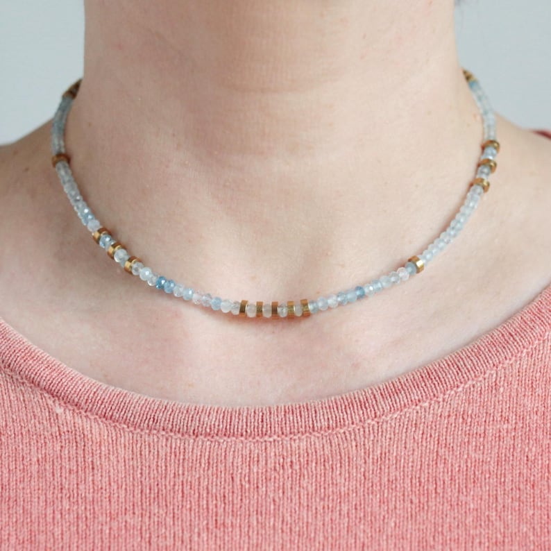 May include: A beaded choker necklace featuring small, translucent light blue faceted beads interspersed with metallic gold-toned cylindrical spacers arranged in a repeating pattern around the entire length of the circular jewelry piece.