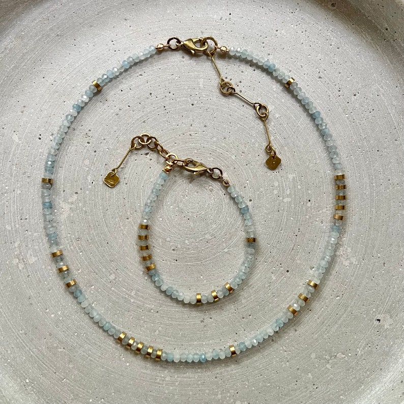 May include: A necklace and bracelet set featuring small, faceted, pale blue beads interspersed with cylindrical gold-toned metal spacers. Both pieces include a gold-toned lobster claw clasp, an adjustable chain extension, and a small square charm.