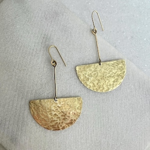 Può includere: Un paio di orecchini color oro. Ogni orecchino presenta un pendente a forma di semicerchio martellato, sospeso da un filo lungo e dritto con un gancio. Gli orecchini hanno una superficie strutturata e una calda lucentezza metallica. Il design è semplice ed elegante.