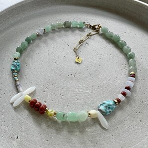 May include: A colourful beaded necklace with green, turquoise, red, and white beads, and gold-coloured accents. The design includes a dragonfly-like element and a gold-toned clasp. The necklace is made with various sizes of beads.