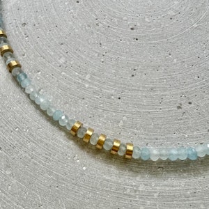 May include: A necklace featuring a strand of small, faceted, pale blue beads interspersed with cylindrical gold-toned metal spacers. The beads have a translucent quality and a reflective surface, arranged in a repeating pattern with the metallic accents.