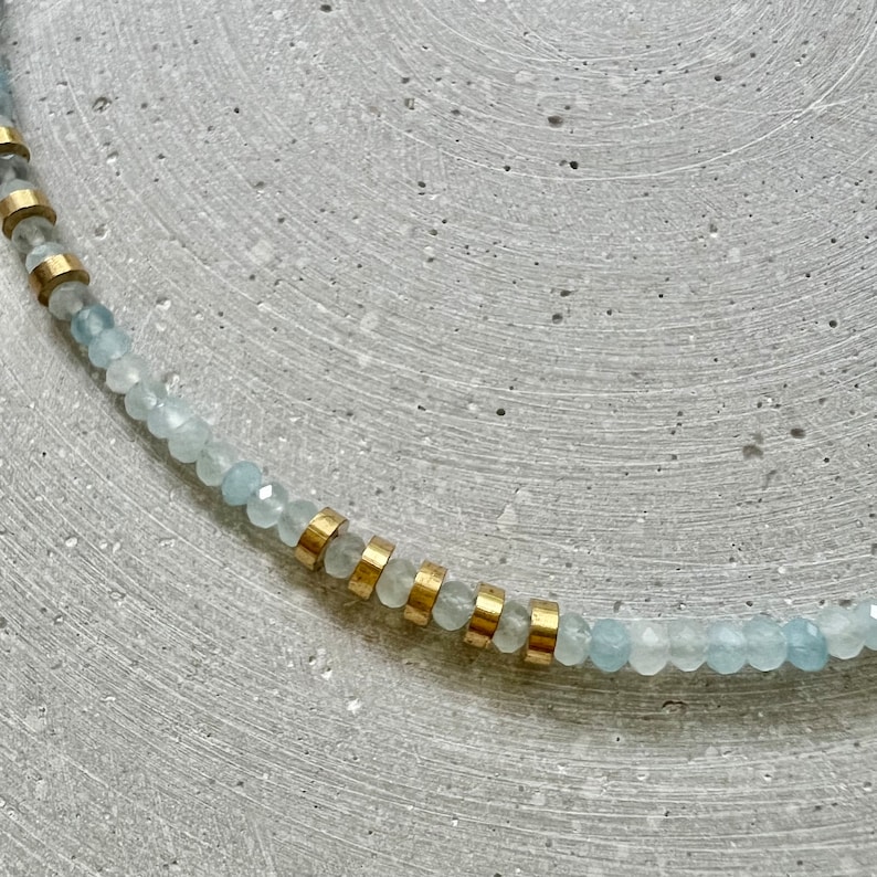 May include: A necklace featuring a strand of small, faceted, pale blue beads interspersed with cylindrical gold-toned metal spacers. The beads have a translucent quality and a reflective surface, arranged in a repeating pattern with the metallic accents.