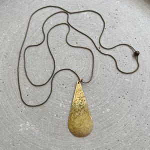 Puede incluir: Un collar largo de latón antiguo con un colgante en forma de lágrima martillada. El colgante es de color dorado cálido y cuelga de una delicada cadena. El collar se muestra sobre una superficie gris.