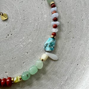 May include: A vibrant gemstone necklace with a mix of colours. The necklace features red, green, blue, and white stones, with gold-coloured metal accents. A large, turquoise-coloured stone is the focal point. The necklace is shown on a grey surface.