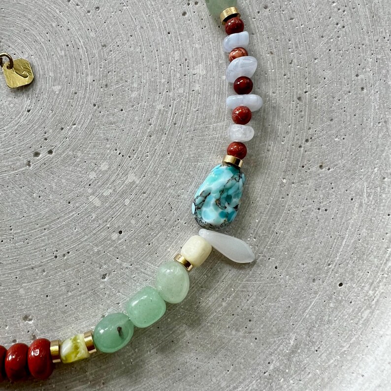 May include: A vibrant gemstone necklace with a mix of colours. The necklace features red, green, blue, and white stones, with gold-coloured metal accents. A large, turquoise-coloured stone is the focal point. The necklace is shown on a grey surface.
