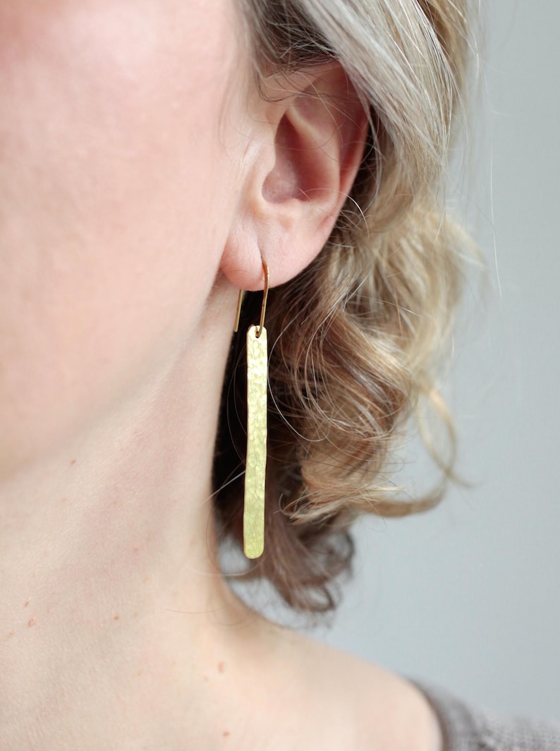 May include: A long, slender, gold-toned earring featuring a flat, rectangular bar with a textured, hammered finish. The earring hangs from a simple, curved hook wire that passes through the earlobe, creating a clean, vertical drop design.
