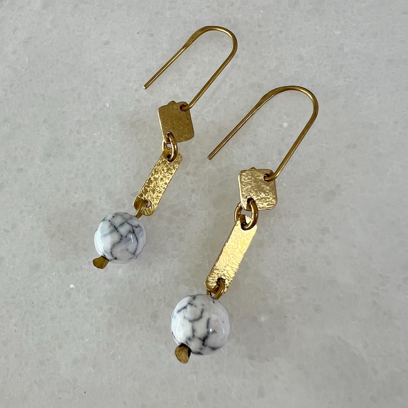 K&ouml;nnte beinhalten: Ein Paar goldene Ohrringe mit wei&szlig;en Marmorperlen und goldenen quadratischen Akzenten. Die Ohrringe sind im H&auml;nge-Stil mit einem einfachen goldenen Haken.