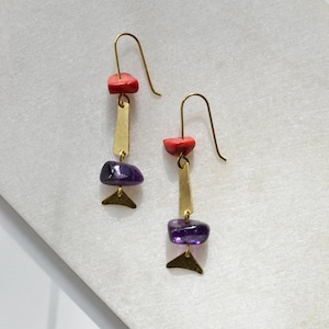 Könnte beinhalten: Ein Paar goldene Ohrringe mit violetten und roten Steinen. Die Ohrringe haben ein geometrisches Design mit einer langen, dünnen Stange und einem kleinen Dreieck am unteren Ende.