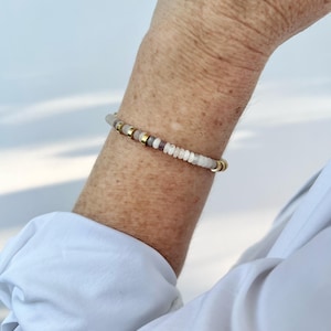 Könnte beinhalten: Ein Perlenarmband bestehend aus einer Abfolge kleiner, runder, durchscheinender weißer und grauer Perlen, die mit dünnen, flachen, metallisch goldfarbenen Zwischenperlen in einem sich wiederholenden Muster um das Handgelenk angeordnet sind.