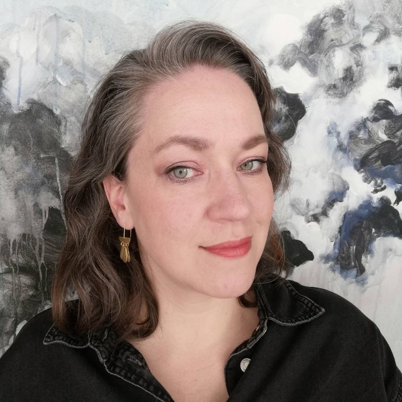 May include: A woman with gray hair wearing a black shirt and gold earrings. She is standing in front of a large abstract painting with shades of gray, white, and blue.