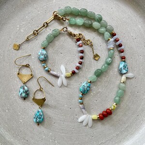 May include: A jewellery set featuring a necklace, bracelet, and earrings. The necklace and bracelet have green, blue, and red beads with gold accents. The earrings have gold-coloured geometric shapes and blue and turquoise teardrop-shaped beads. The set includes a dragonfly charm.