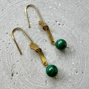 K&ouml;nnte beinhalten: Ein Paar goldene Ohrringe mit einem gr&uuml;nen Malachit-Stein an jedem Ohrring. Die Ohrringe haben ein schlichtes, geometrisches Design.