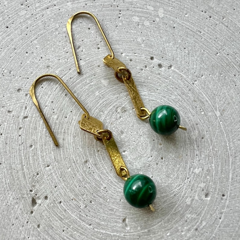 K&ouml;nnte beinhalten: Ein Paar goldene Ohrringe mit einem gr&uuml;nen Malachit-Stein an jedem Ohrring. Die Ohrringe haben ein schlichtes, geometrisches Design.