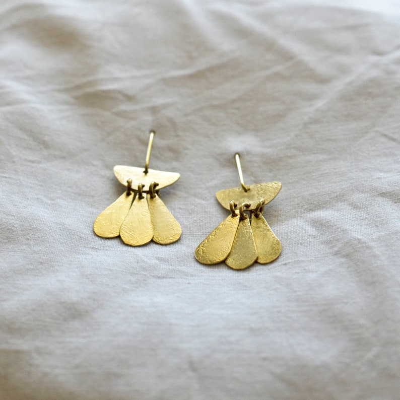 May include: A pair of gold earrings with a geometric design. Each earring features a small, curved top piece with three teardrop-shaped pieces hanging below.