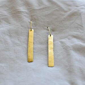 May include: A pair of gold-toned drop earrings featuring long, thin, rectangular metal bars with a hammered, textured finish. Each bar is suspended from a simple, curved wire hook, creating a clean and elongated silhouette for these dangling earrings.