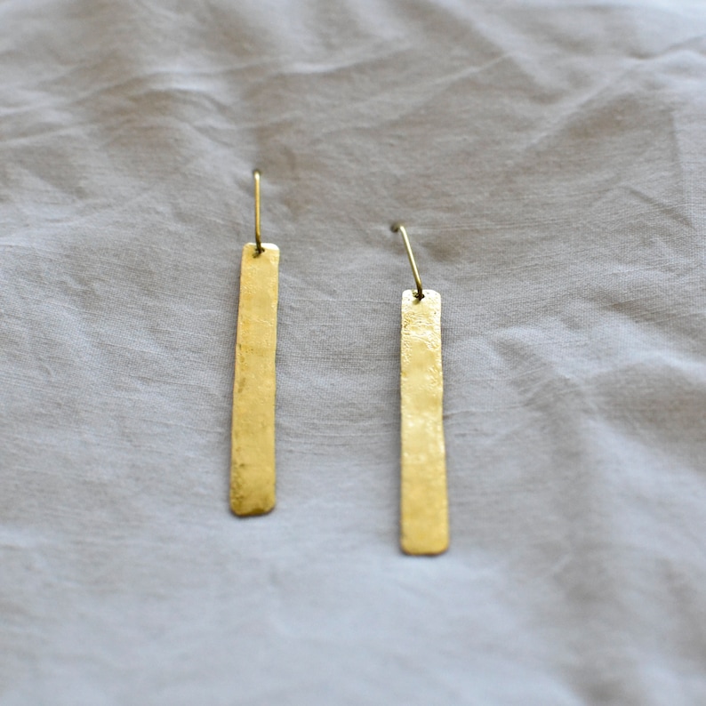 May include: A pair of gold-toned drop earrings featuring long, thin, rectangular metal bars with a hammered, textured finish. Each bar is suspended from a simple, curved wire hook, creating a clean and elongated silhouette for these dangling earrings.