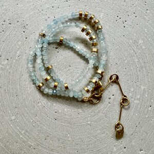May include: A multi-strand necklace featuring faceted pale blue beads interspersed with small cylindrical gold-toned metal spacers. The necklace includes a gold-toned metal lobster claw clasp and an adjustable extender chain with a small metal tag.