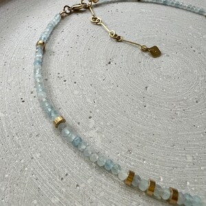 May include: A necklace featuring small, faceted, pale blue beads interspersed with cylindrical gold-toned metal spacers. The piece includes a gold-toned metal chain extender with a small, flat, rectangular tag attached to the end of the clasp mechanism.