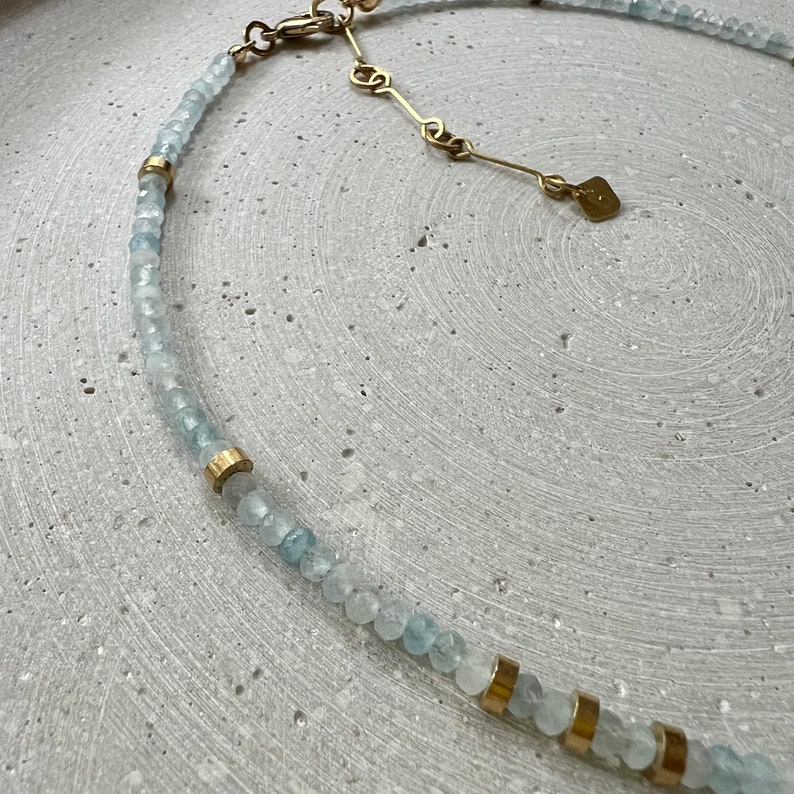 May include: A necklace featuring small, faceted, pale blue beads interspersed with cylindrical gold-toned metal spacers. The piece includes a gold-toned metal chain extender with a small, flat, rectangular tag attached to the end of the clasp mechanism.