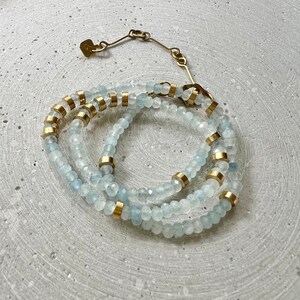 May include: A multi-strand bracelet featuring faceted pale blue beads interspersed with cylindrical gold-toned metal spacers. The bracelet includes a gold-toned metal chain extender with a small square tag at the end, secured by a lobster clasp.