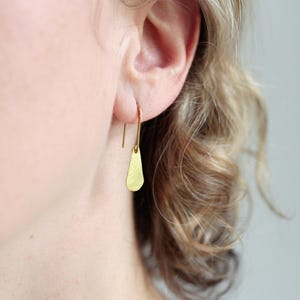 Könnte beinhalten: Ein goldfarbener Ohrring mit einem langen, schlanken Haken, der durch das Ohrläppchen geführt wird und mit einem kleinen, flachen, tropfenförmigen Anhänger mit texturierter, matter Oberfläche verbunden ist, der vertikal am Haken hängt.