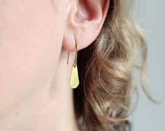 May include: A gold-toned earring featuring a long, slender hook wire that passes through the earlobe, connected to a small, flat, teardrop-shaped pendant with a textured, matte surface finish that hangs vertically from the base of the wire.