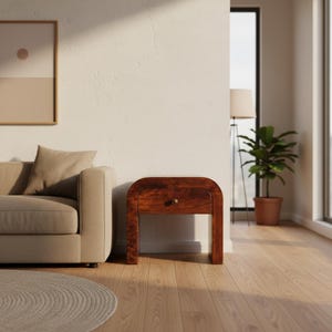 May include: A wooden nightstand with a single drawer and a curved top. The nightstand is a rich brown color and has a brass knob. The nightstand is in a room with a sofa, a rug, and a potted plant.