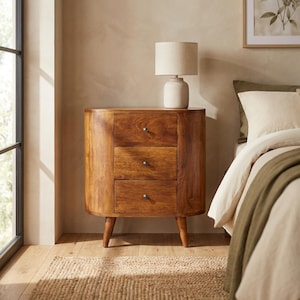 May include: A three-drawer wooden nightstand with a warm brown finish. The nightstand has a curved front and sits on four tapered legs. A cream-colored lamp with a cylindrical shade rests on top. The nightstand is next to a bed with white bedding.