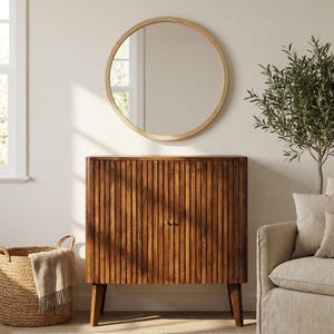 Könnte beinhalten: Ein Holzschrank mit vertikalen Rillen und kleinen goldenen Knöpfen. Darüber ein runder Spiegel mit hellem Holzrahmen. Ein geflochtener Korb und ein kleiner Baum sind ebenfalls im Bild.
