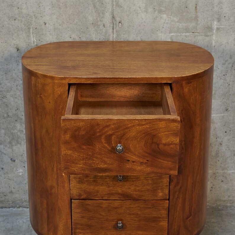 Może przedstawiać: Drewniana szafka nocna z trzema szufladami, z kt&oacute;rych jedna jest otwarta. Szafka nocna ma ciepły brązowy kolor i zaokrąglony kształt. Uchwyty szuflad są małe i metalowe.
