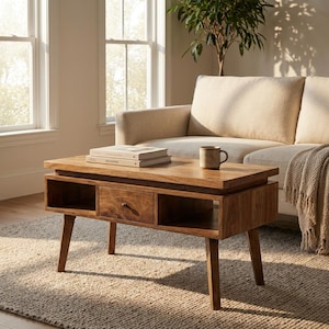 May include: A wooden coffee table with a drawer and open storage. The table has a natural wood finish and is supported by four angled legs. Books and a mug are on top. The table is approx. 120 cm long.