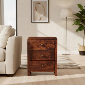 May include: A three-drawer wooden nightstand with a warm, brown finish. The nightstand has rounded edges and brass-colored knobs. The drawers are evenly spaced. The nightstand is a piece of bedroom furniture.