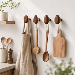 May include: Wooden kitchen accessories hanging on a white wall. Includes a beige apron, a wooden brush, a wooden spoon, and a small cutting board. The items are suspended from dark brown wooden wall hooks. A neutral-toned kitchen setting.