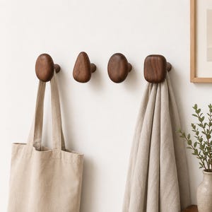 May include: Four dark brown wooden wall hooks with varying organic shapes are mounted on a white wall. A beige tote bag hangs from the first hook, and a beige towel hangs from the fourth hook. A framed picture and a vase with greenery are in the background.