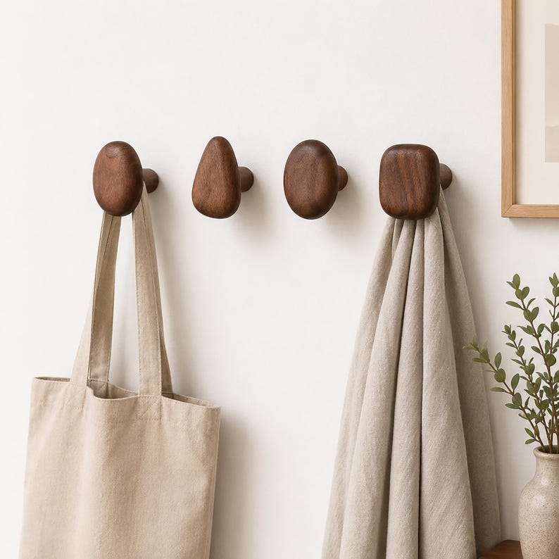 May include: Four dark brown wooden wall hooks with varying organic shapes are mounted on a white wall. A beige tote bag hangs from the first hook, and a beige towel hangs from the fourth hook. A framed picture and a vase with greenery are in the background.