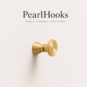 Op de afbeelding: Een goudkleurige metalen wandhaak met een uitlopende voet en een platte, ronde voorkant. Boven de haak staat de tekst PearlHooks, gevolgd door de woorden SIMPLE. MODERN. FUNCTIONAL. De haak is bevestigd op een effen, lichtgekleurde ondergrond.