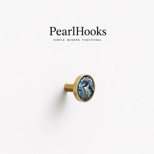 May include: A round wall hook with a brushed brass finish and a circular inlay of iridescent blue and green abalone shell. The text PearlHooks SIMPLE. MODERN. FUNCTIONAL. is printed above the hook in a black serif and sans-serif font.