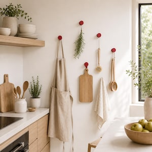 Op de afbeelding: Een keukeninterieur met een beige schort, houten snijplanken en keukengerei dat aan rode haken hangt. Een houten plank toont witte kommen en borden. Groene kruiden en planten voegen een natuurlijk tintje toe. Een kom met groene appels.