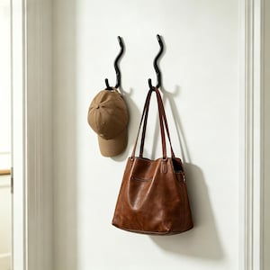 May include: Two black metal wall hooks in a wavy design. A brown baseball cap hangs from one hook, and a brown leather tote bag hangs from the other. The items are against a white wall.