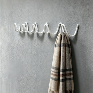 Könnte beinhalten: Ein weißer Wand-Kleiderhaken aus Metall mit einem einzigartigen, wellenförmigen Design. Ein beiger, brauner und schwarzer Schal mit Karomuster hängt an einem der Haken. Der Haken ist an einer strukturierten grauen Wand befestigt.