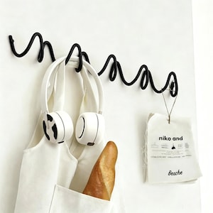 May include: A black metal wall hook with a unique, wavy design. White headphones hang from the hook, along with a white tote bag holding a loaf of bread. A small tag with the text "niko and Beache" is also hanging.