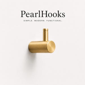 Op de afbeelding: Een cilindrische wandhaak van messing met een geborstelde afwerking en een kleine verticale pin. De tekst PearlHooks SIMPLE. MODERN. FUNCTIONAL. staat boven de haak. Het ontwerp heeft een horizontale basis en een korte, opstaande paal.