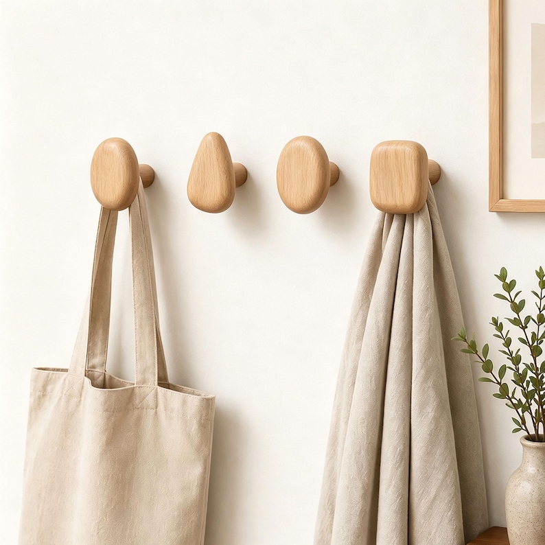 May include: Four light-colored wooden wall hooks with smooth, rounded geometric shapes. One hook holds a beige canvas tote bag, and another hook holds a folded light gray linen cloth. The hooks are mounted horizontally in a row on a plain wall.